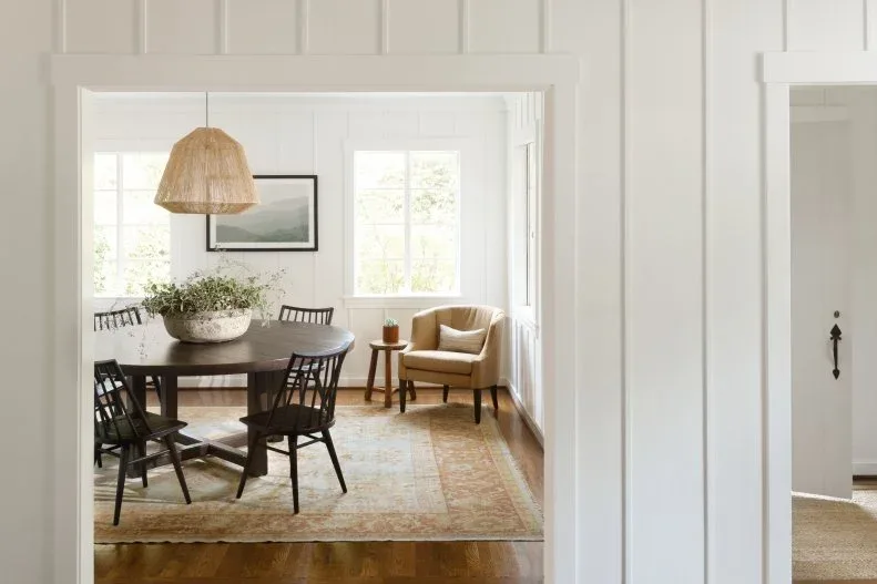 Round dark dining table with black chairs, a woven pendant and a pale armchair near windows
