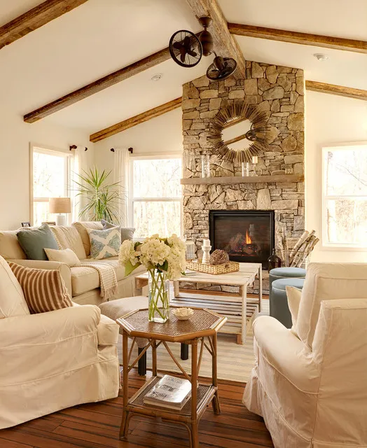 Pale chairs and a sofa gather around a substantial stone fireplace beneath timber beams.