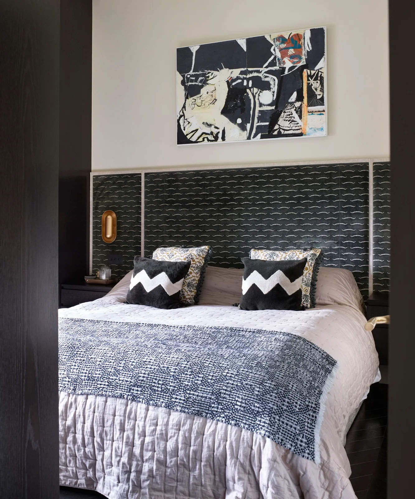 A dark patterned headboard wall sits beneath a framed graphic picture above black-and-white zigzag pillows and pale bedding with a fine dark repeat