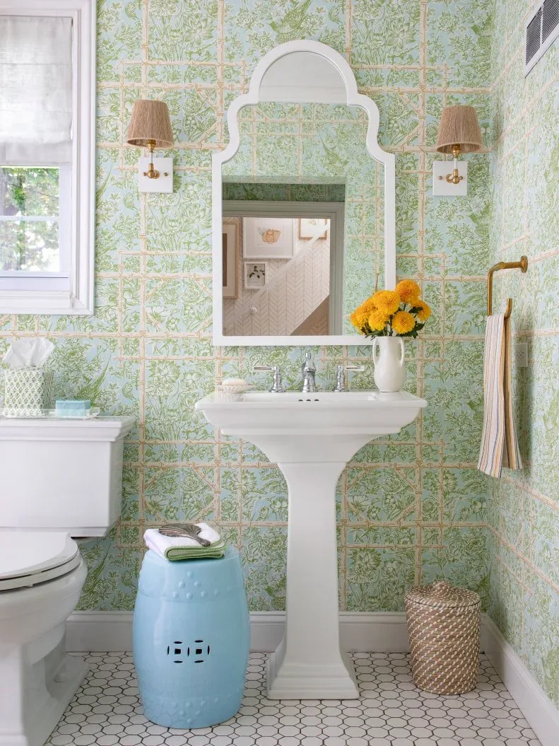 Powder room with pale green botanical wallpaper, white shaped mirror and pedestal basin, woven light shades, light blue stool and small woven basket.