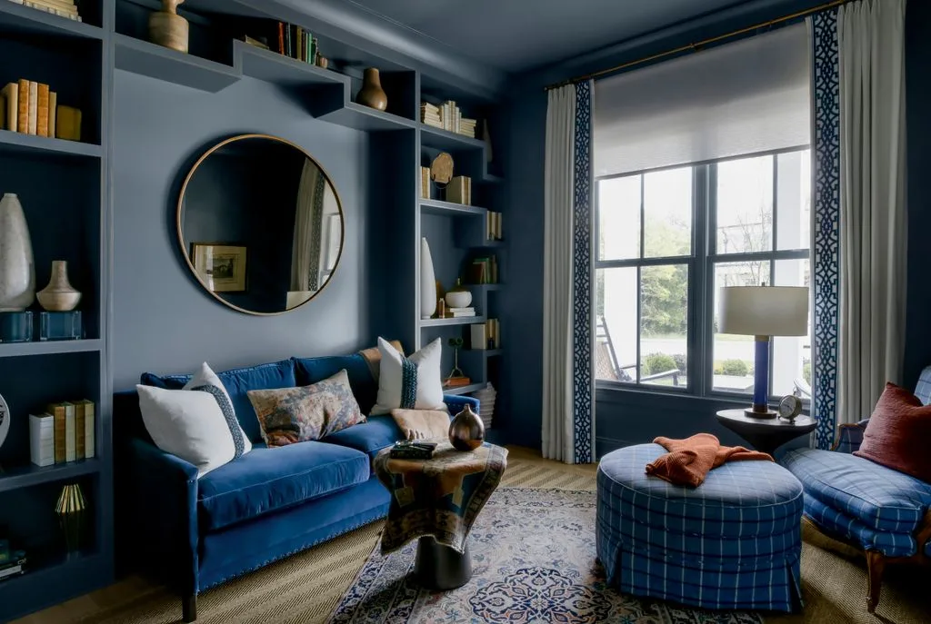 Dark blue bedroom sitting area with built-in shelves, a blue sofa, round mirror, plaid ottoman, and a window.