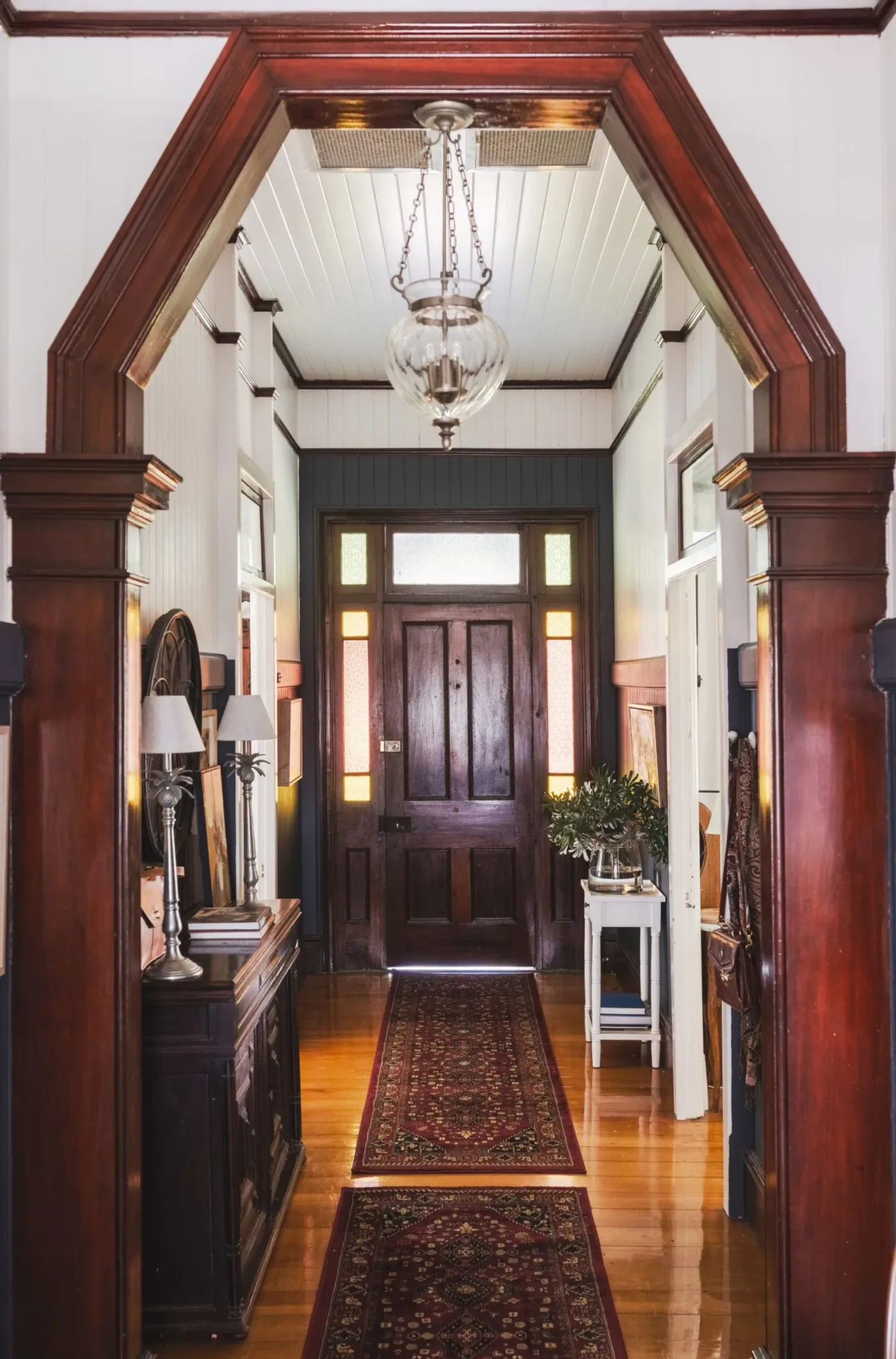 Long entrance hallway with dark wooden door and decorative timber arch, pale walls, patterned runners and a narrow dark cabinet.