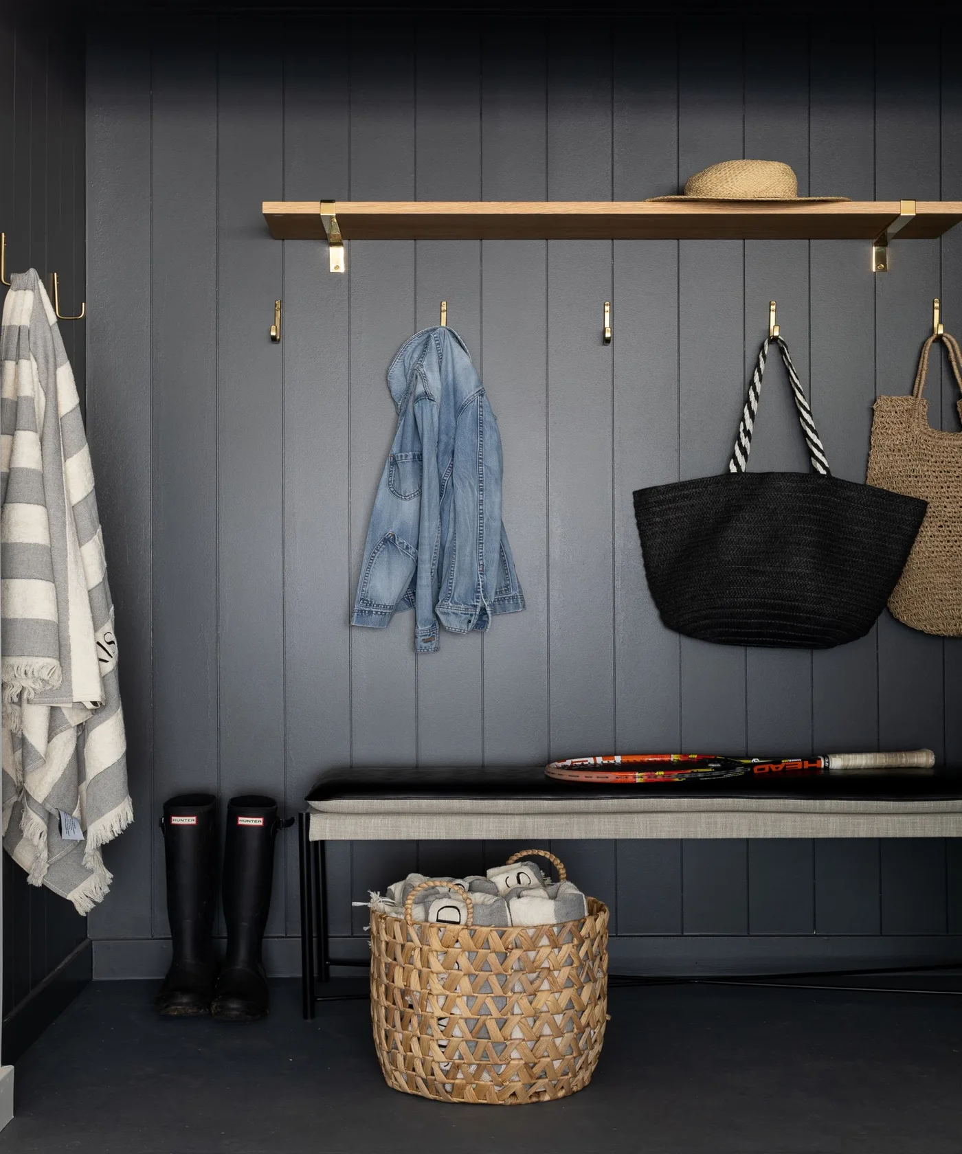 Dark gray paneled entry with wood shelf, small wall hooks, narrow bench, hanging bags, tall boots and a woven basket