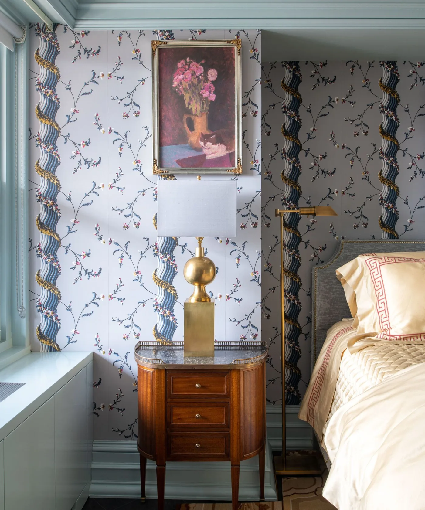 Delicate flowering wallpaper beside pale blue window trim, a gold lamp and framed flower artwork.