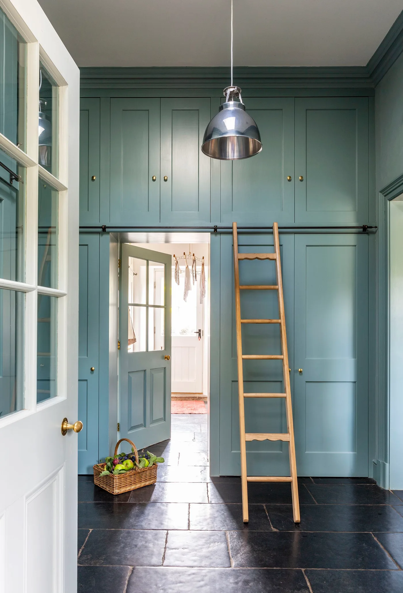 Muted teal hallway with paneled cupboards around a doorway, overhead cabinets and a wooden access ladder.
