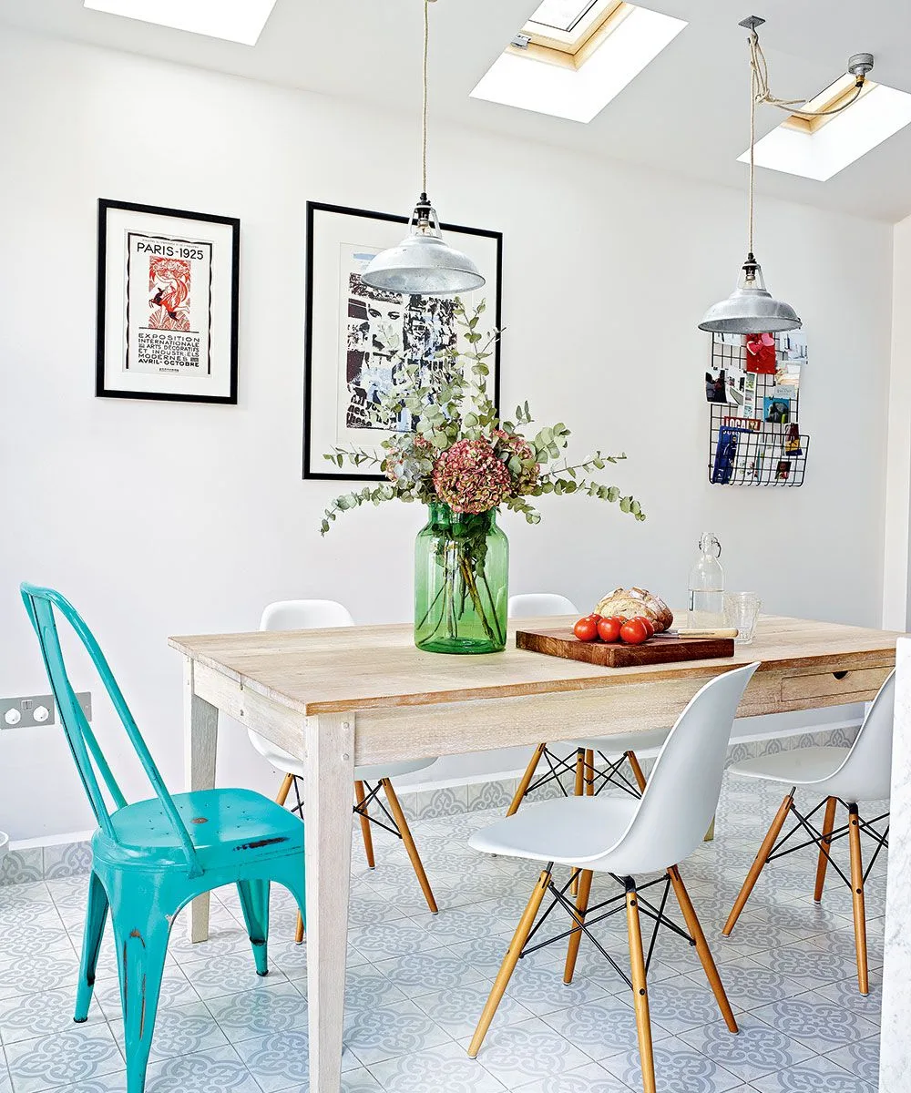 Two pale metal pendants over a wood table with white chairs and a turquoise chair beneath skylights