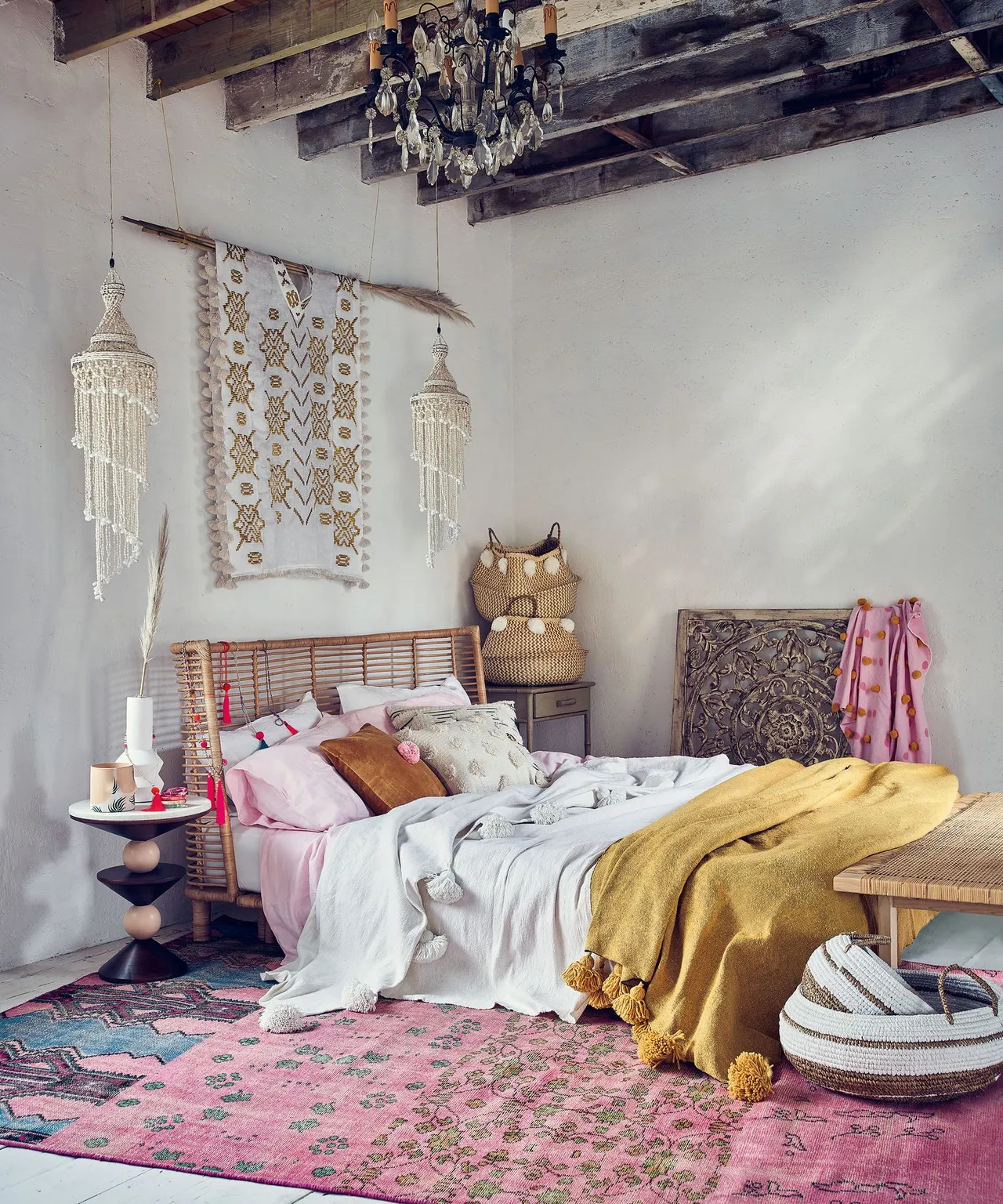 Bedroom with woven bed, pink and white bedding, yellow throw, patterned pink rug, exposed beams, hanging fabric decoration and chandelier.