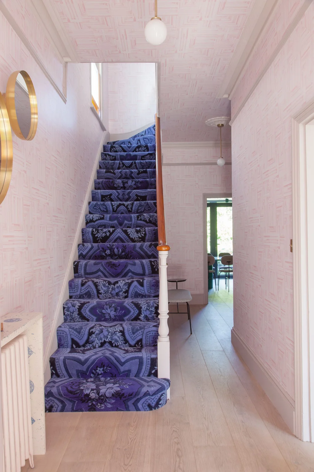 Pale pink patterned walls and ceiling surround a blue floral stair runner, gold-colored oval mirrors and small white globe lights