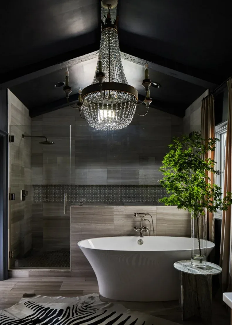 Bathroom with charcoal vaulted ceiling, hanging chandelier, gray shower walls, white freestanding bath, clear shower screen, and large plant beside a window.