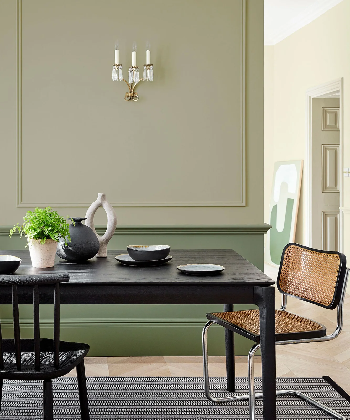 A dark wooden table and woven-seat chair sit beside two muted green wall colors, pale vessels, dark bowls and a small plant