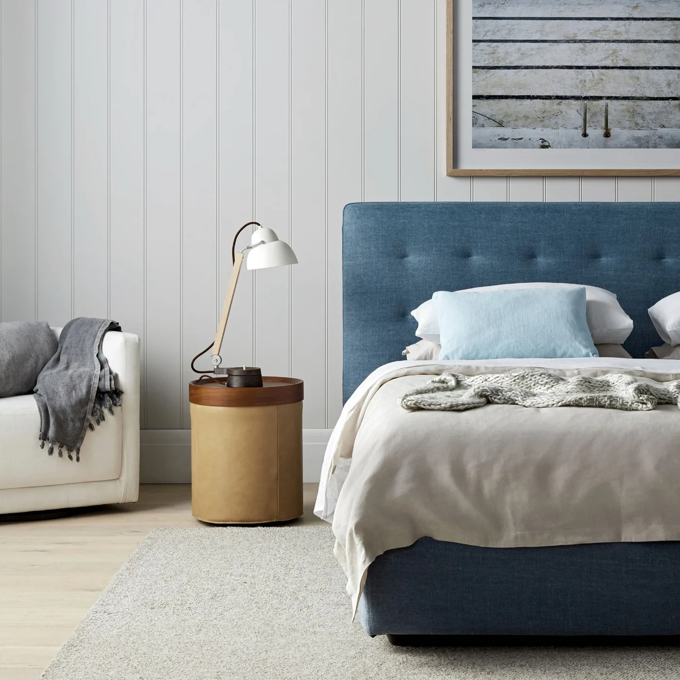 Bedroom with a blue upholstered bed, pale vertical wall paneling, round warm-toned bedside table and pale bedding.