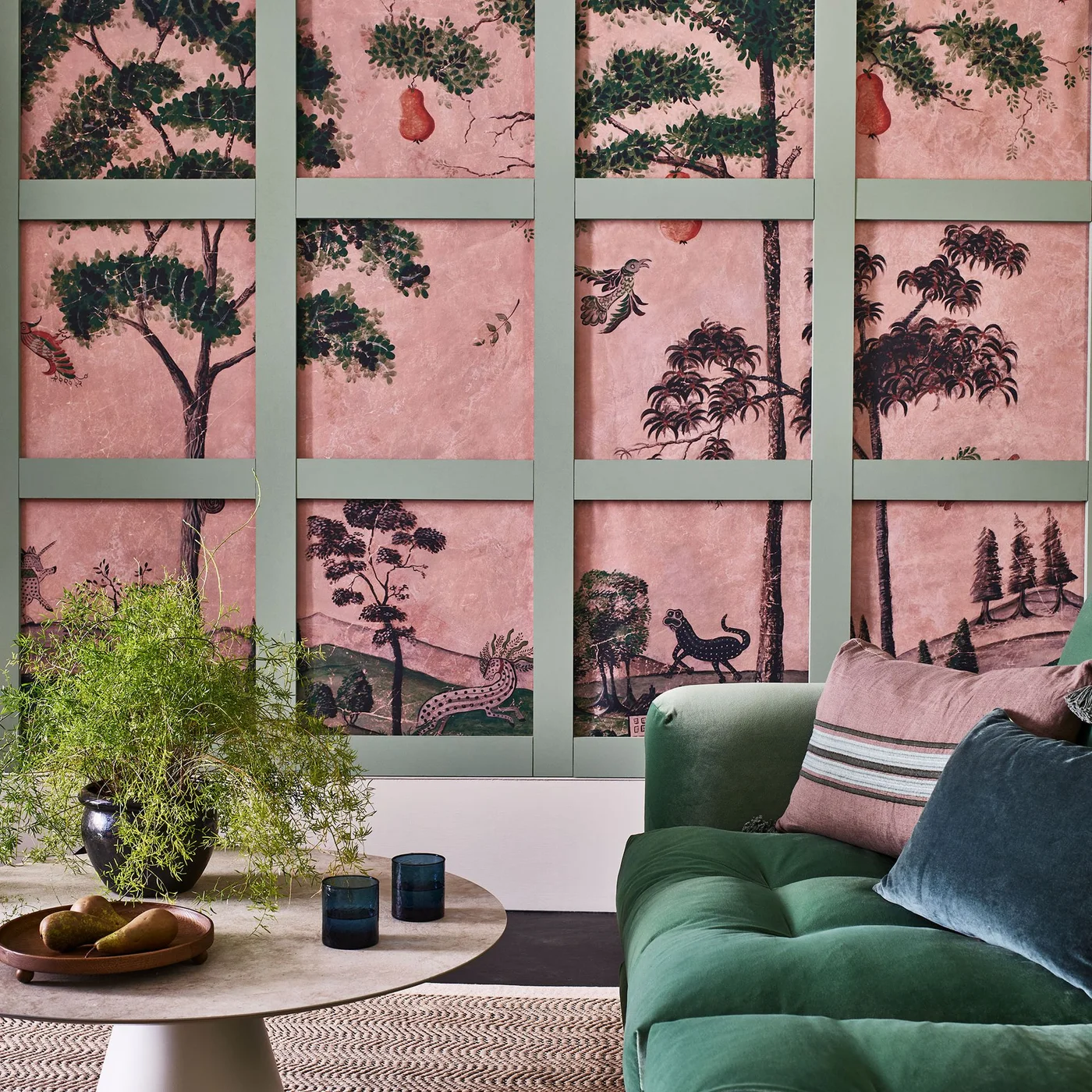 Green framing divides a pink woodland mural into rectangles behind a green sofa and round pale coffee table