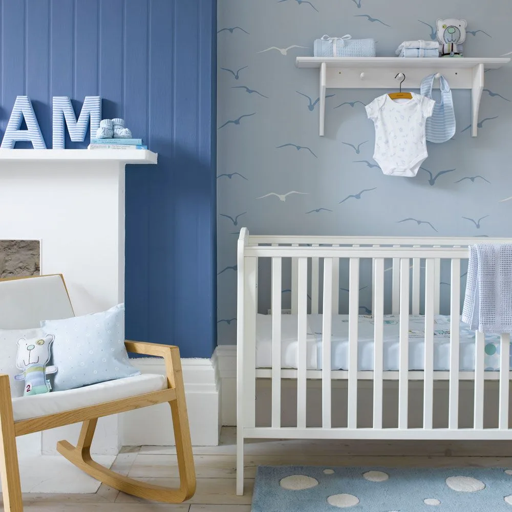 White crib and wooden rocking chair against pale bird wallpaper and deeper blue paneling beside lighter fireplace surround.