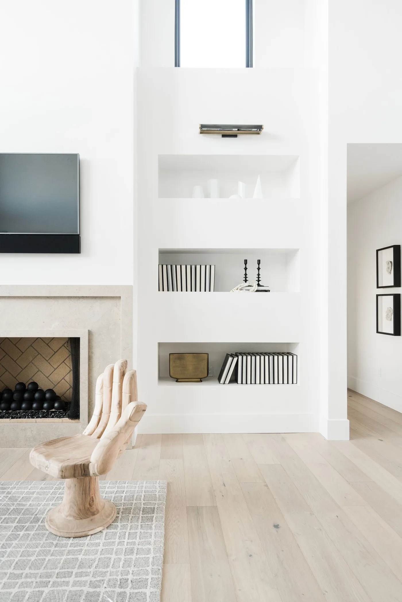 A carved hand-shaped wood chair stands beside a pale fireplace wall with simple inset shelving, dark-framed artwork and a light wood floor