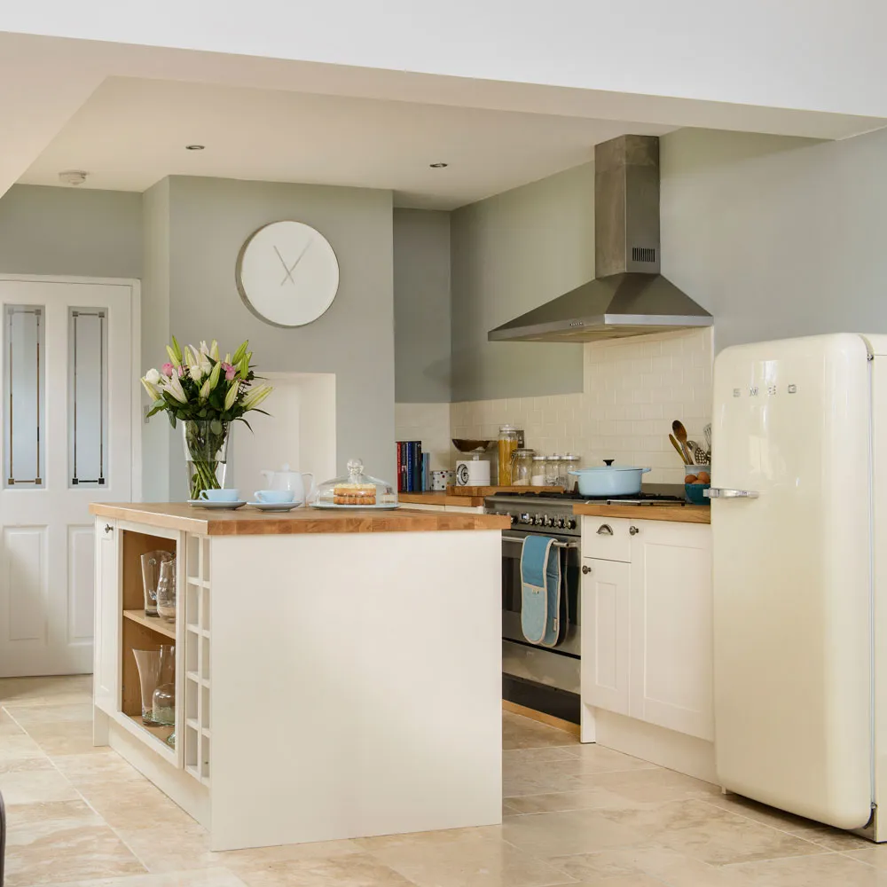 Cream lower cabinets and a small wooden-topped island sit below muted green walls beside a white refrigerator
