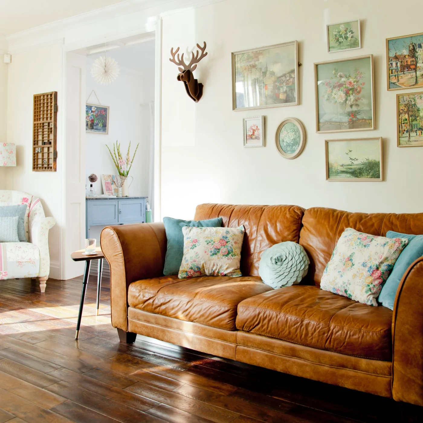 A tan sofa with floral cushions sits below a varied gallery of framed pictures on a pale wall