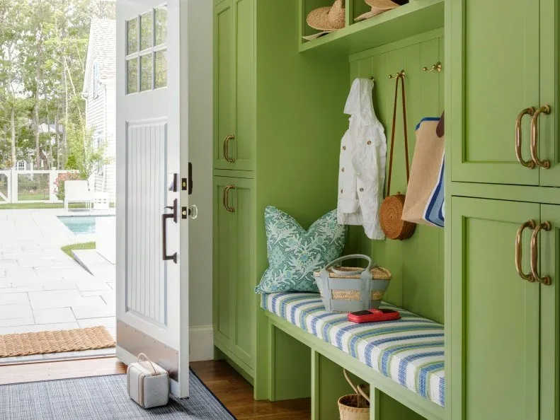Green paneled entry storage with striped bench cushion, lower cubbies, hanging bags and tall side cupboards beside white door