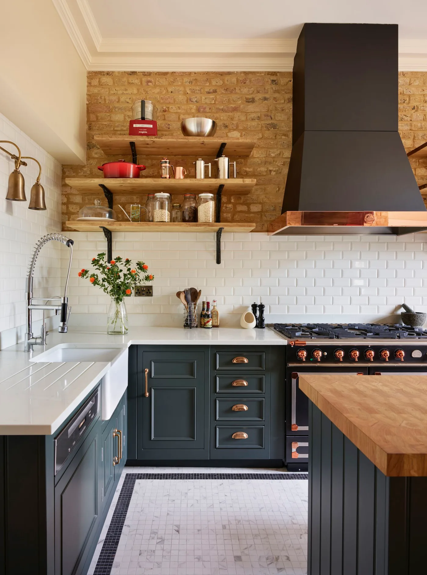 Cottage kitchen with dark green cabinets, white sink and rectangular backsplash tiles, exposed brick above, wooden shelves and a dark hood with warm metal trim.