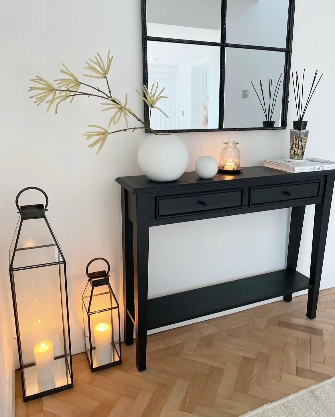 A slim dark console with two drawers stands beneath a black grid mirror beside pale vessels and two floor lanterns on a wooden floor