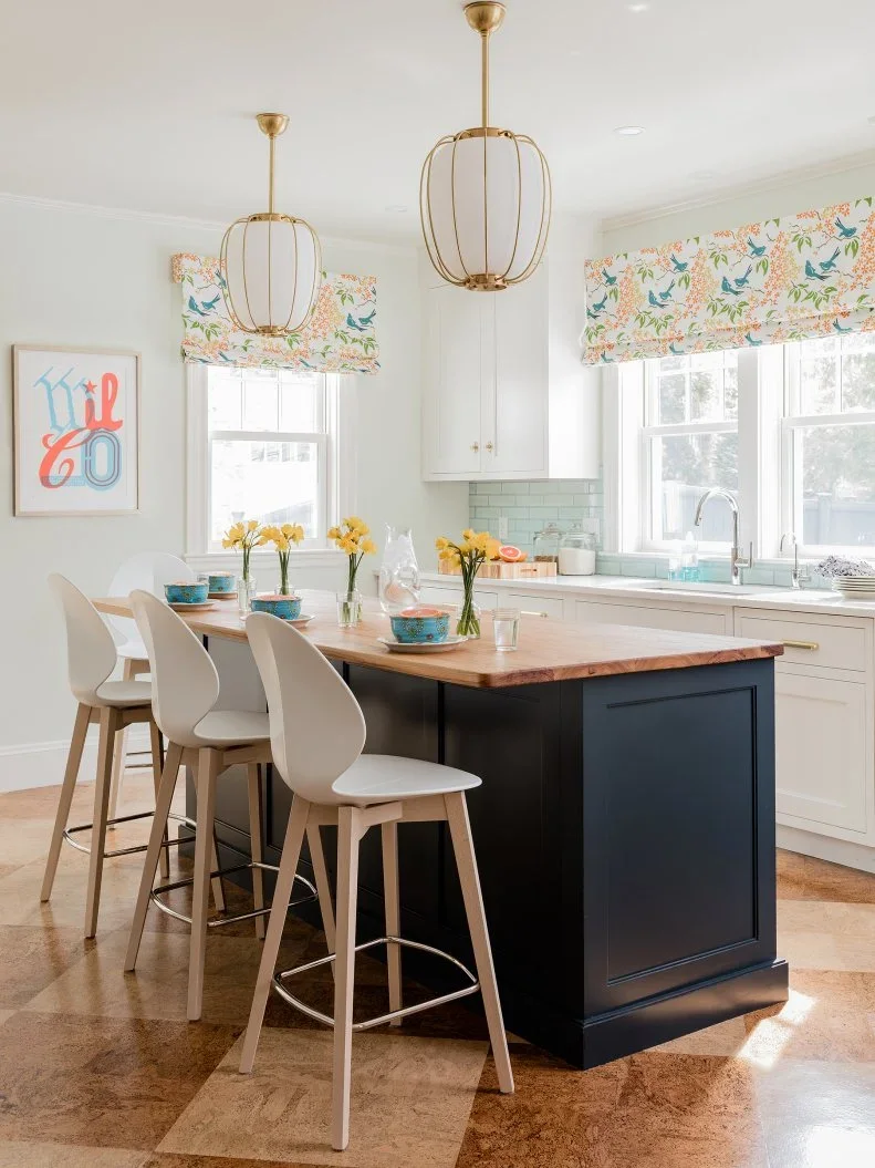 A dark paneled island with a wood top stands beside pale cupboards, white curved stools, warm-toned pendant lights and small colorful window shades