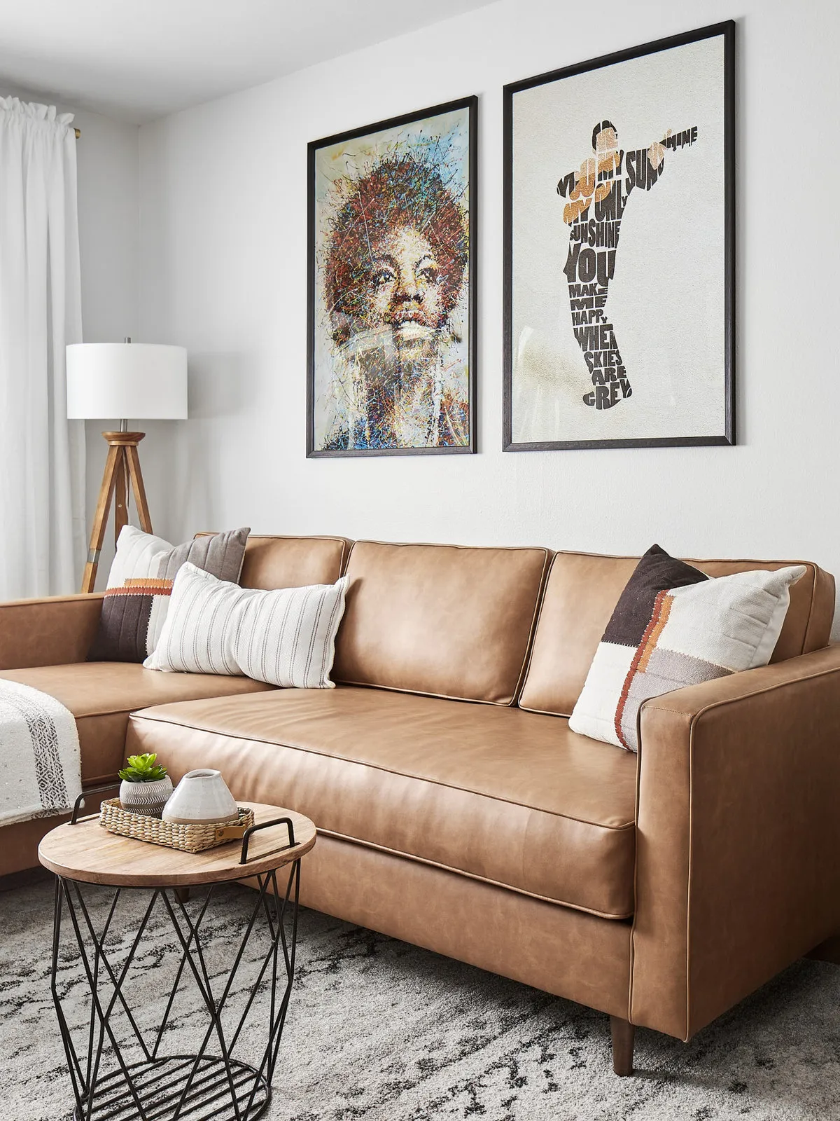 A tan leather sofa stands beneath framed artwork beside a small wire-based table.