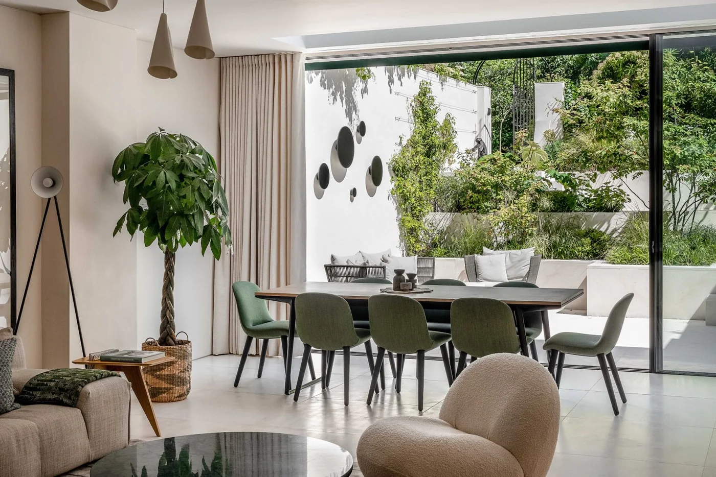 Muted green chairs surround a long pale table beneath narrow cone-shaped pendants beside tall curtains, a leafy plant and broad garden glazing