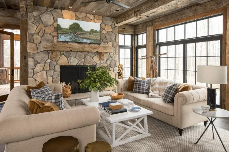 Full-height irregular stone fireplace beneath weathered beams, with landscape artwork, pale sofas and checked cushions