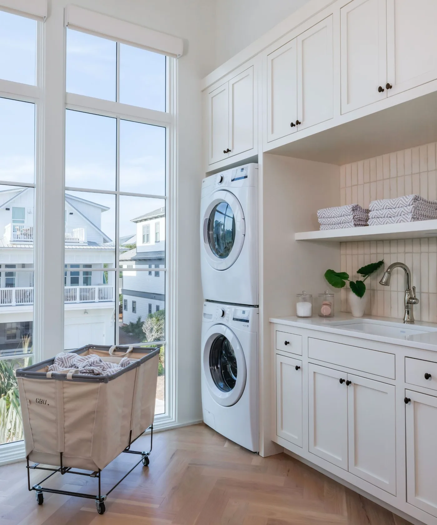 White laundry cabinets with stacked machines, paneled sink recess, folded towels and wheeled fabric hamper beside tall windows