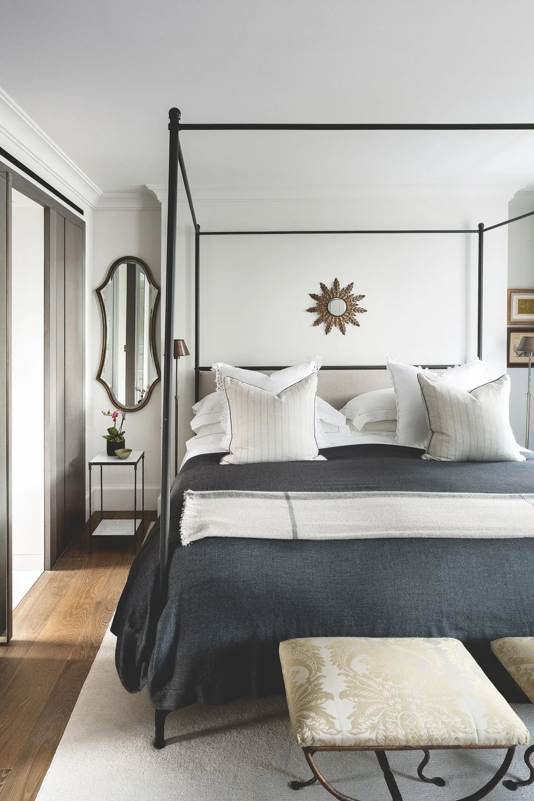 A slender dark four-poster bed stands in a white room with dark bedding, pale pillows, wall mirrors and a patterned footstool above a light rug