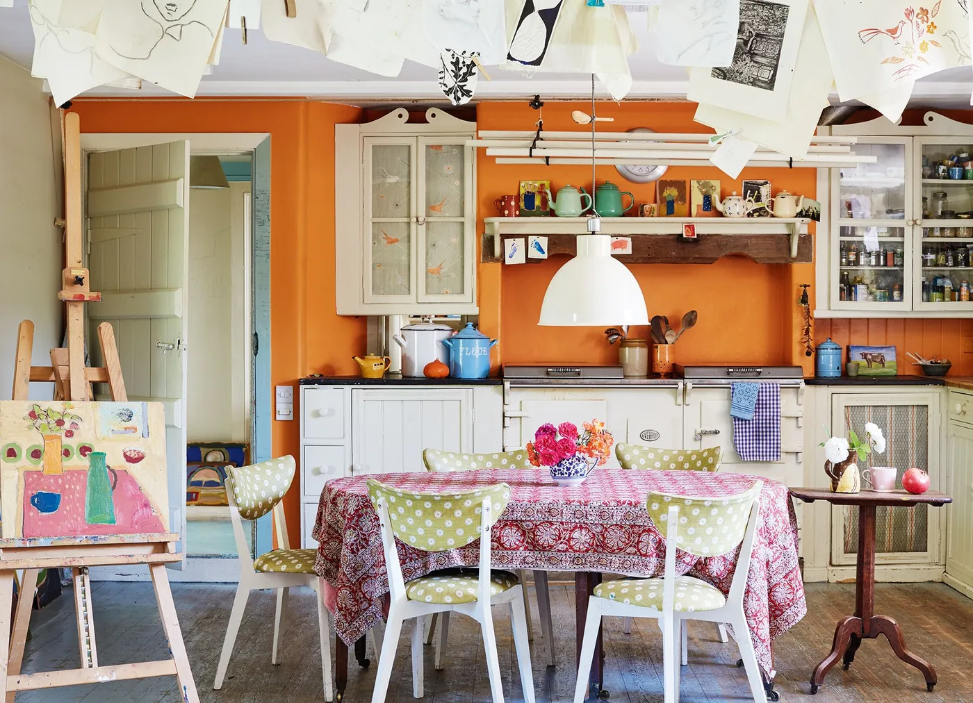 Orange-walled kitchen with a pink patterned tablecloth, green spotted chairs and pale cabinets.
