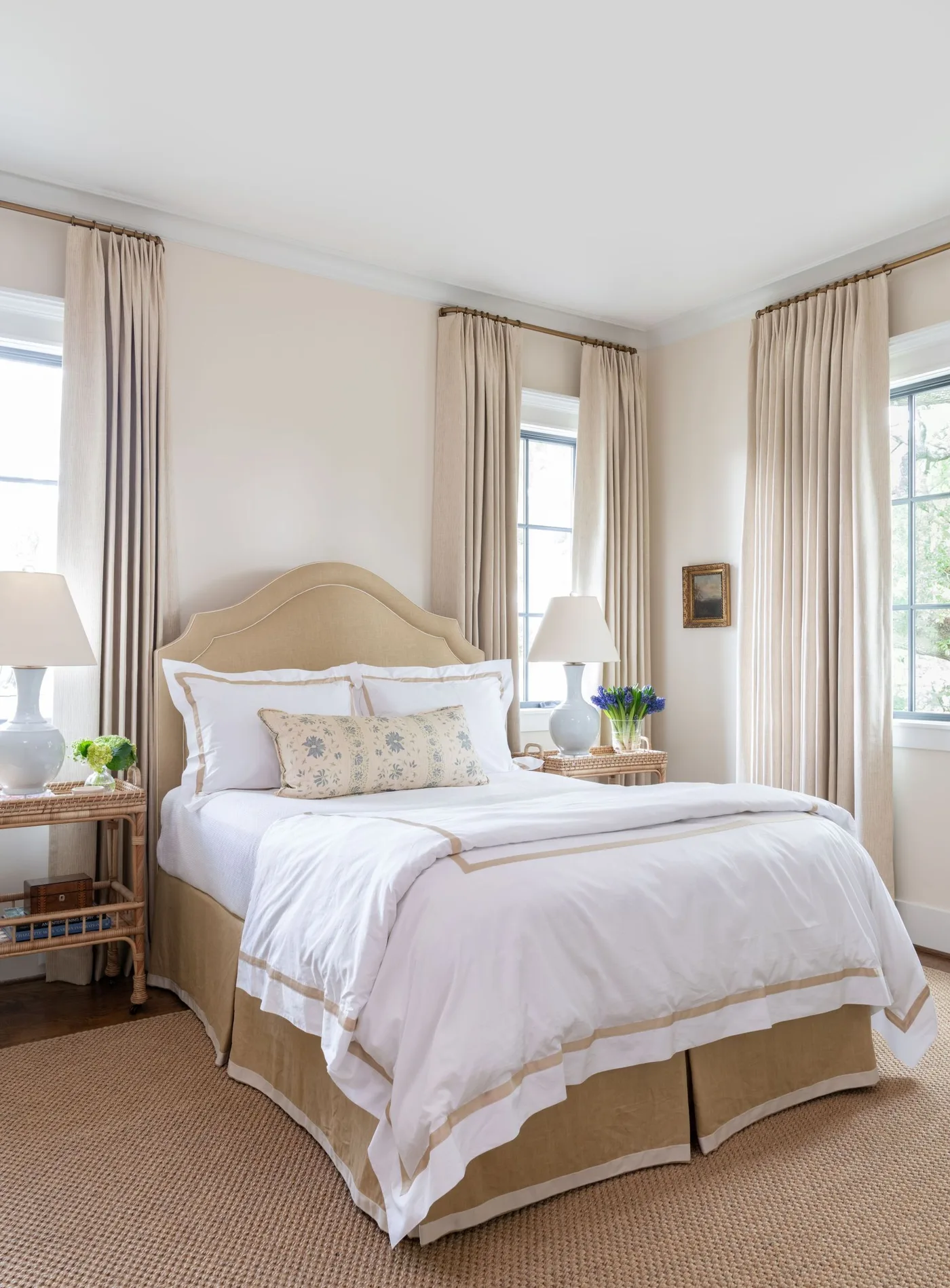 Pale bedroom with a curved beige headboard, white bedding, woven bedside tables and long neutral curtains.