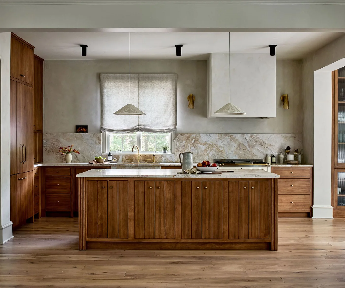 A wooden island and matching cabinets sit beneath pale pleated pendants beside a broad veined backsplash, pale hood and window blind