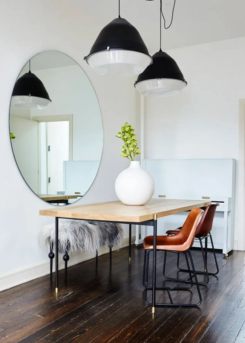 Tall oval mirror beside a narrow wood dining table with brown chairs, a textured bench and black pendants