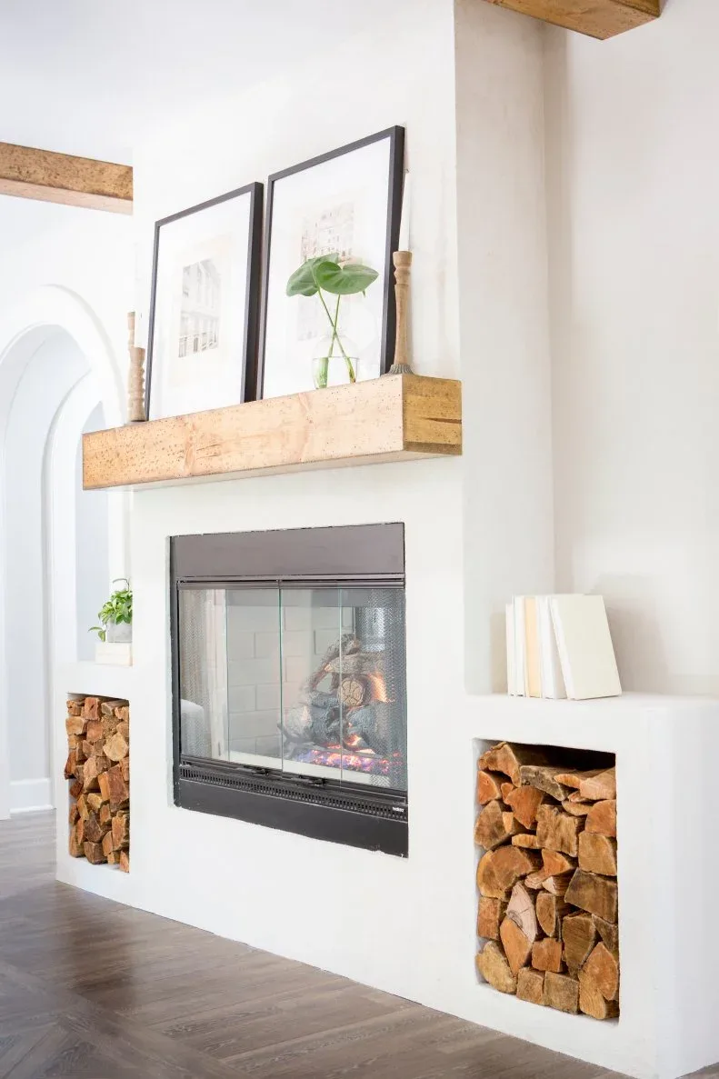 White fireplace wall with thick wood mantel, black-framed glazed firebox, two framed pictures and lower log-filled niches