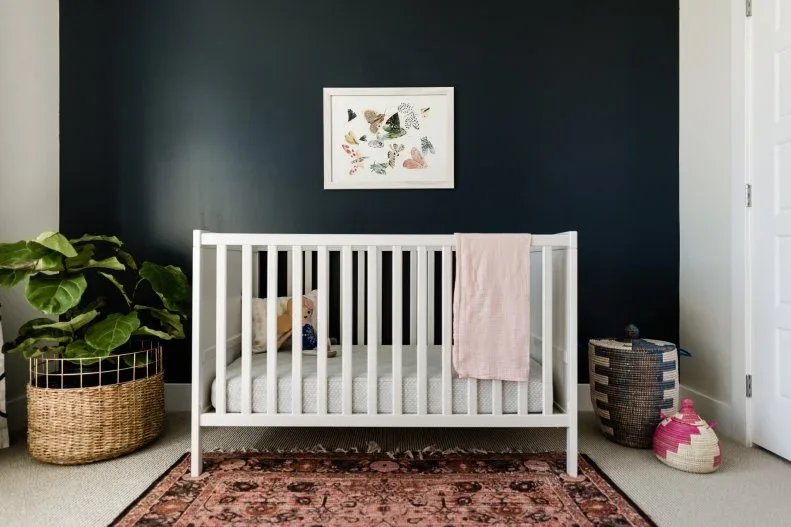White crib against dark wall with botanical picture, warm patterned rug, woven containers, plant and draped pink blanket.