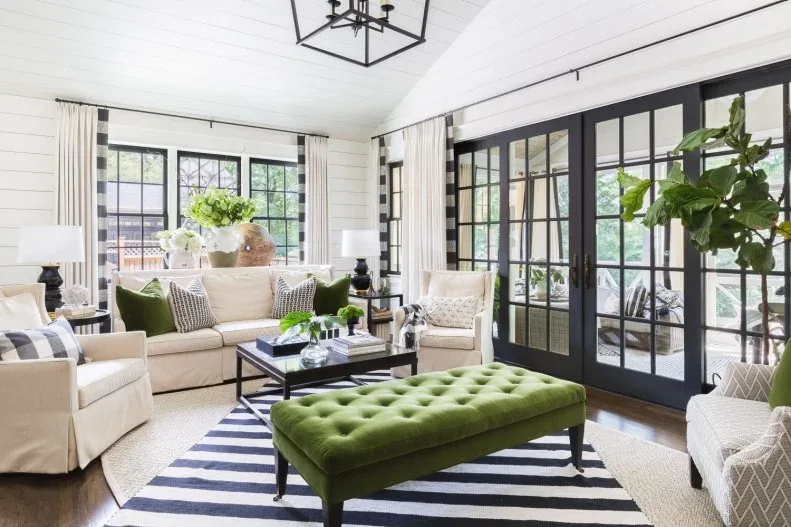 White board-walled room with black-framed glazed doors, pale sofas, green tufted bench and black-and-white striped rug