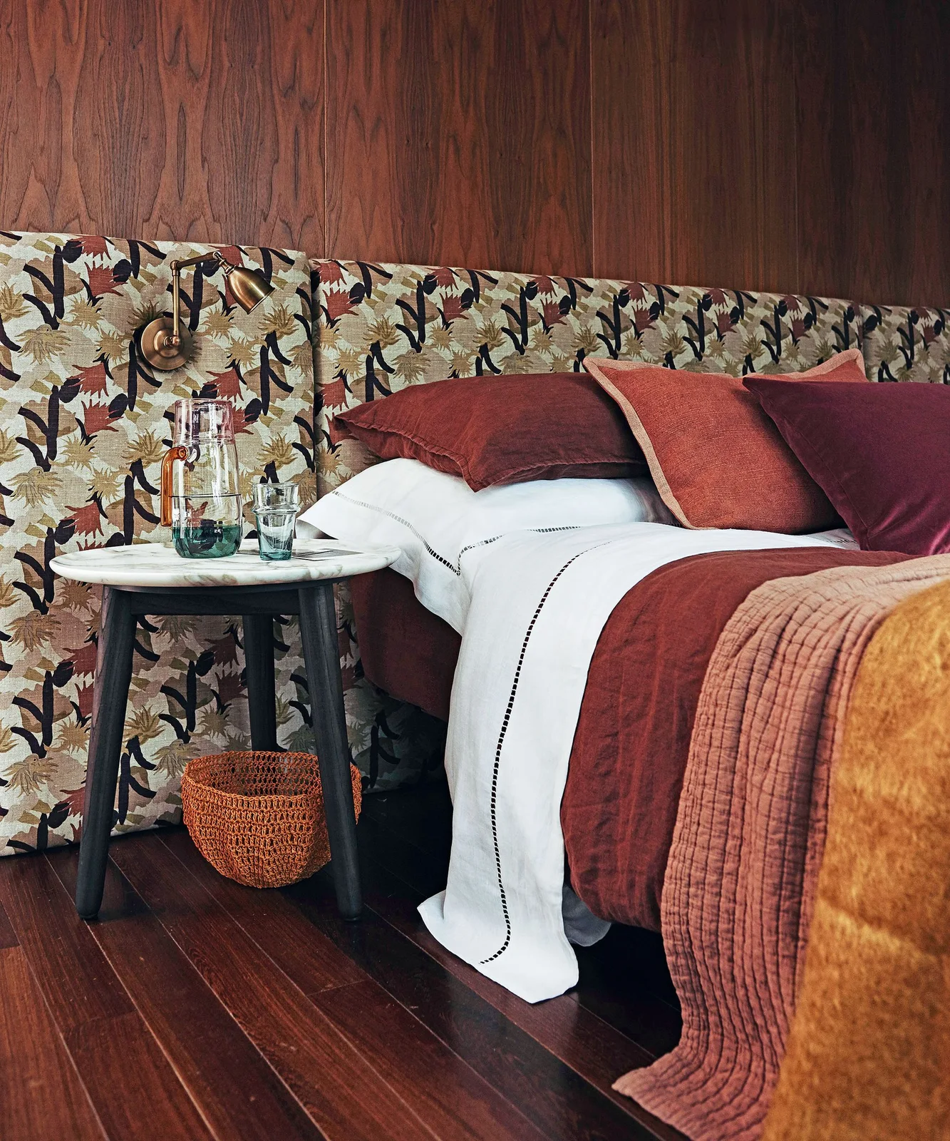 A broad floral upholstered headboard sits against reddish wood-grain walls behind pale bedding and rust-colored pillows