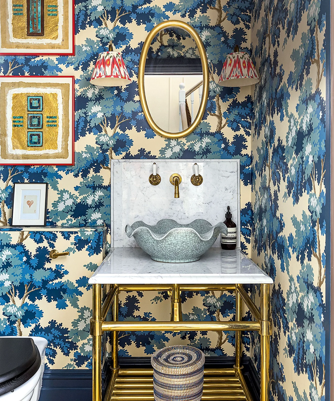 Blue botanical wallpaper surrounds a scalloped basin beneath an oval gold-toned mirror and red patterned wall-light shades