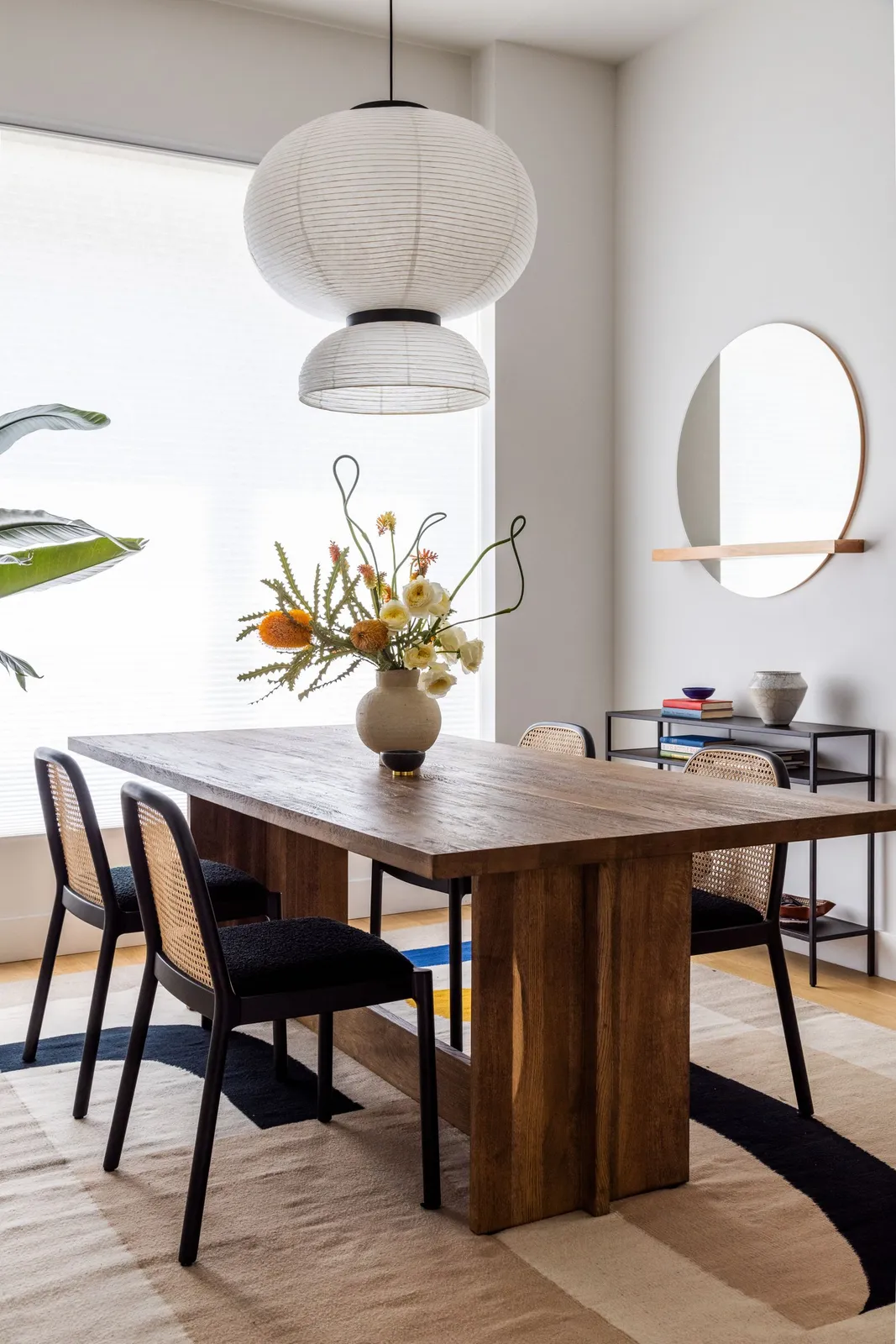 A broad wooden dining table sits beneath a large pale pendant beside dark cane-backed chairs and an oval wall mirror