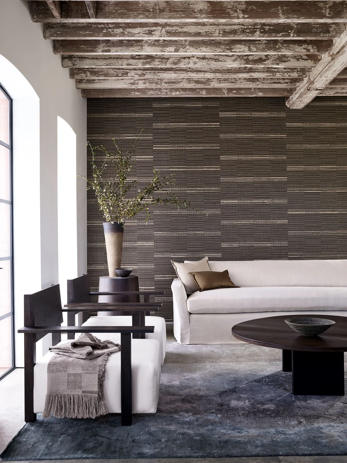 A pale sofa and wood-framed chairs sit against a dark horizontally patterned wall beneath a textured wood ceiling beside a round dark table