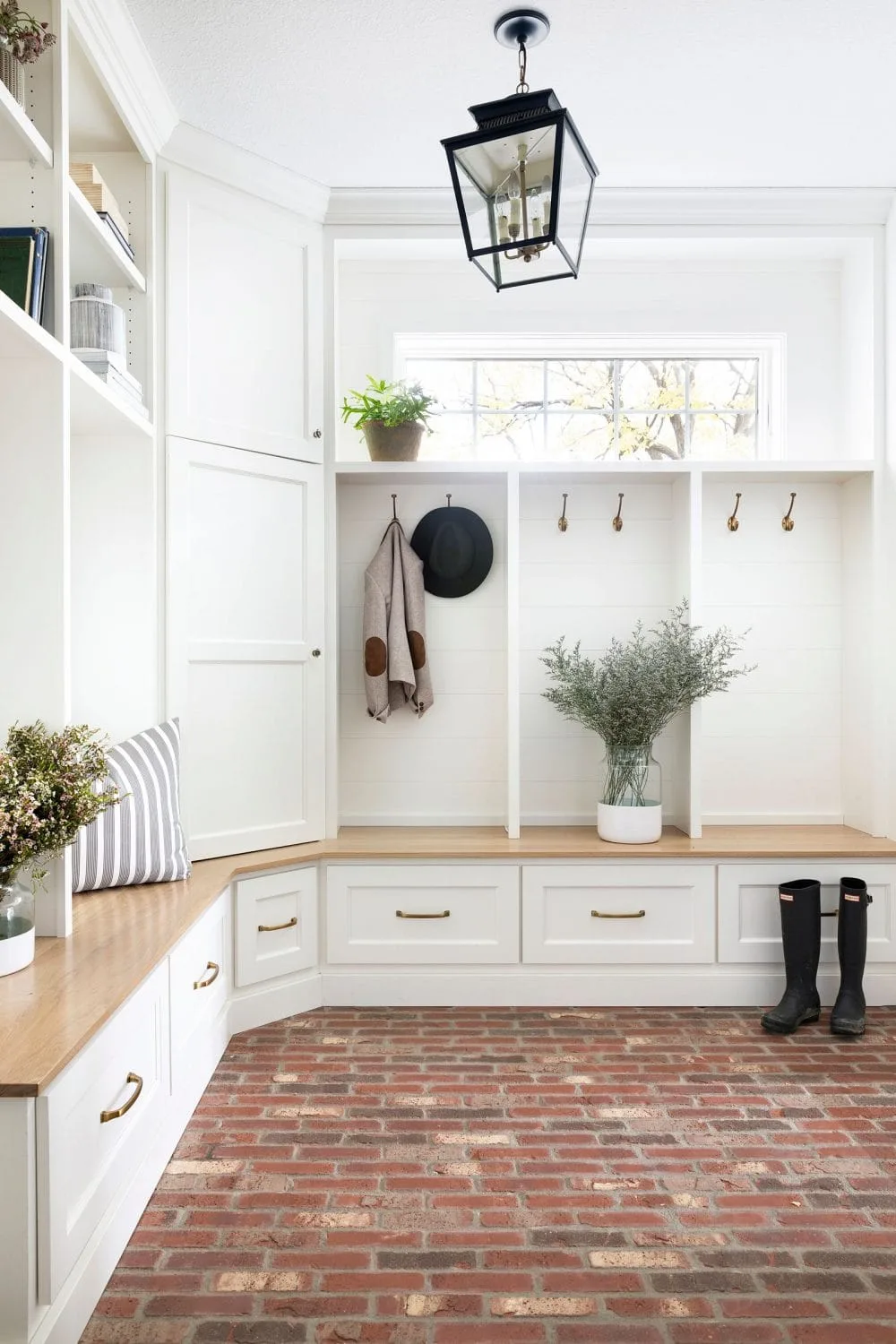White L-shaped mudroom bench with wooden seats, hooks, a black lantern and a red brick-toned floor.