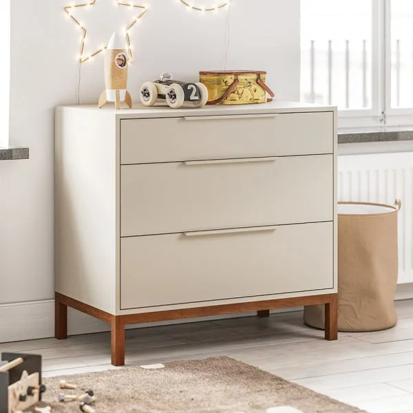 A pale three-drawer chest with wooden legs and simple pulls stands between two windows beside a fabric storage basket