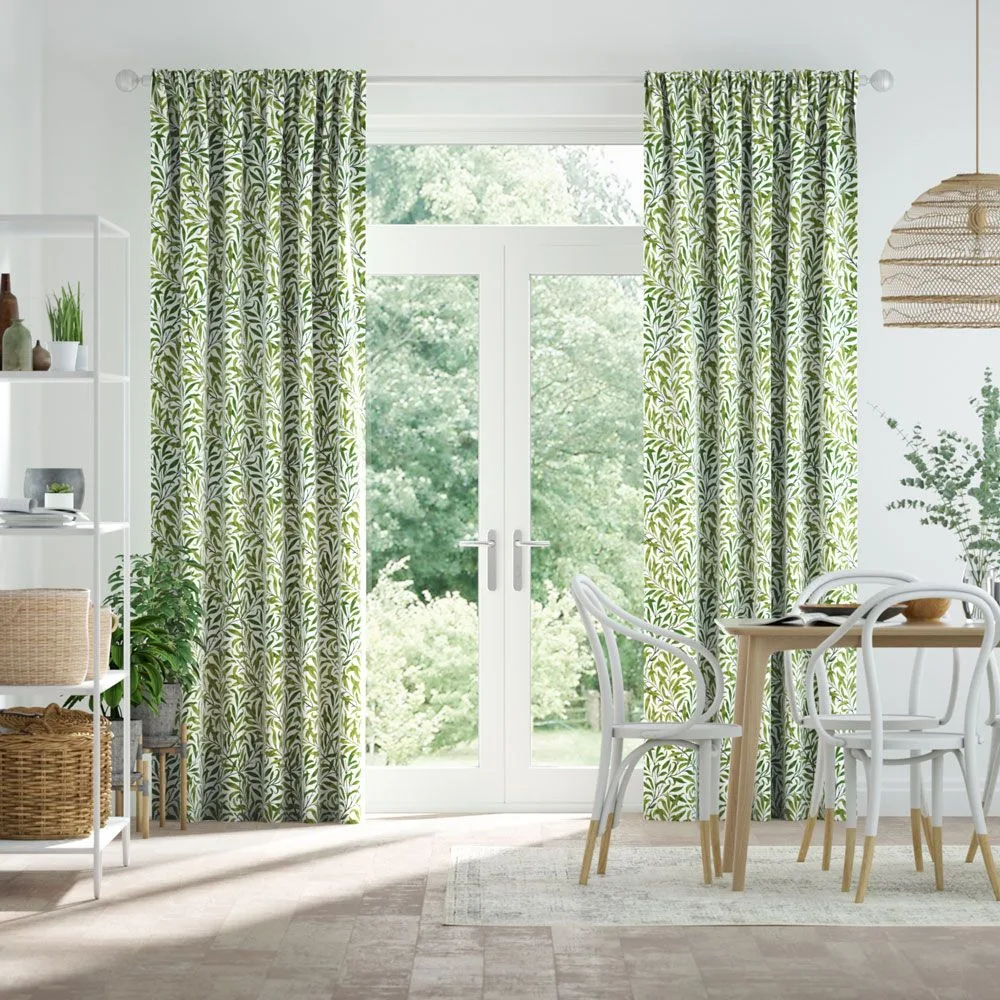 Green-and-white leaf-patterned curtains around pale glazed garden doors, beside a wooden dining table, white chairs, woven light shade, and open shelving.