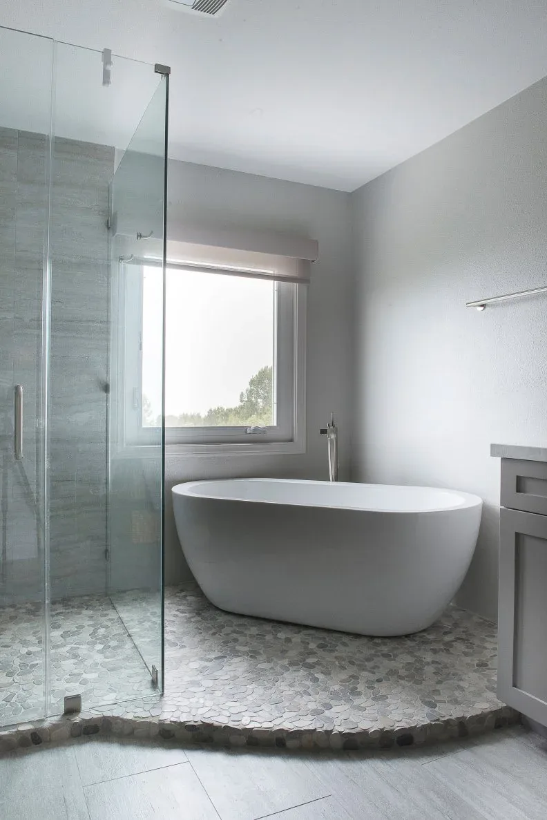 Pale freestanding bath on a raised rounded-mosaic platform beside a glass shower and gray walls