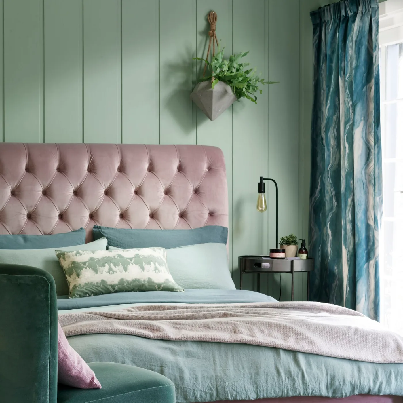 Muted green paneled bedroom with a pale rose tufted headboard, green bedding, patterned curtains and a dark round table