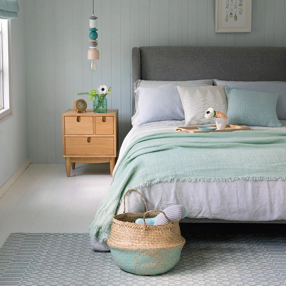 A gray upholstered headboard sits behind pale bedding and a mint throw beside wooden drawers, a small pendant and a woven basket