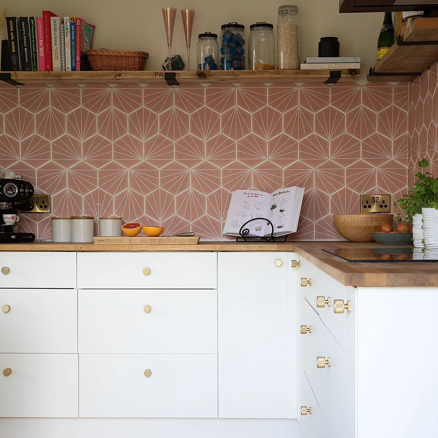 Pink geometric tiles wrap a kitchen corner beneath wooden shelves beside white drawers and wooden counters