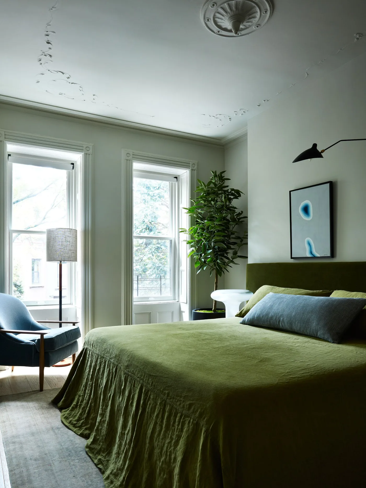 Light bedroom with olive bedding and a wide low green headboard, a teal chair, a large plant and a black reading light