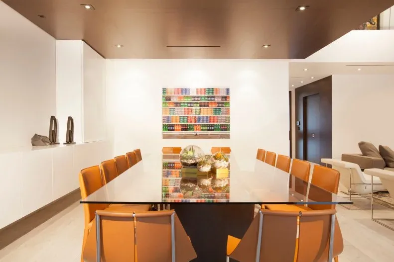 Orange dining chairs with slim metal frames along a glass table in a pale contemporary room, with colorful rectangular artwork behind.