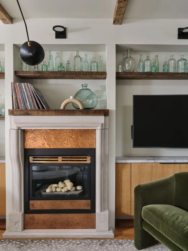 Small fireplace with pale surround and warm-toned inner panel beneath wood shelves of glass bottles beside a television